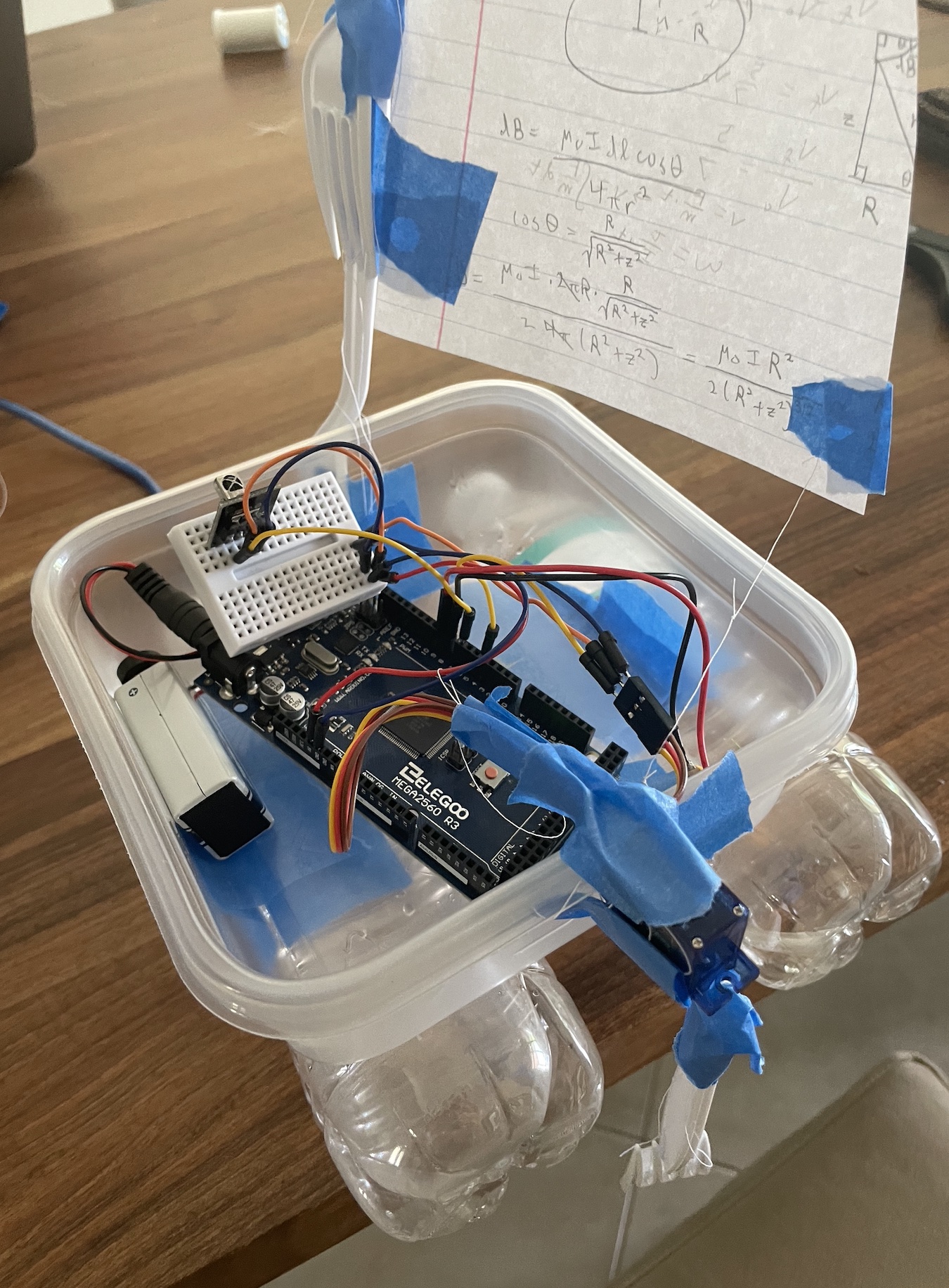 A plastic box containing a circuit, supported by two plastic bottles. Two plastic forks are taped together in the front and connect to a paper sail with physics equations on it.